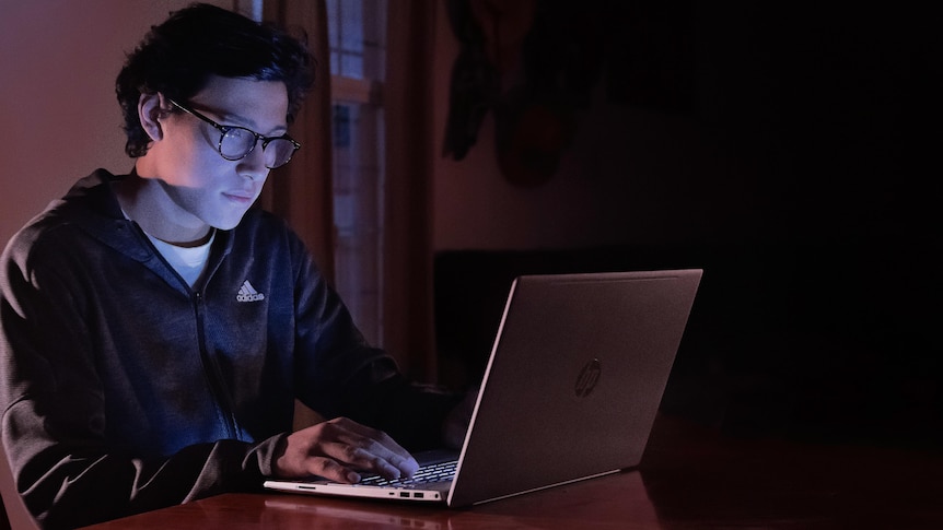 A man earing glasses works on a laptop in a dark room.