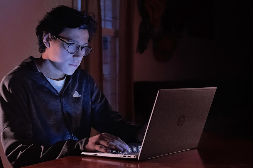 A man earing glasses works on a laptop in a dark room.
