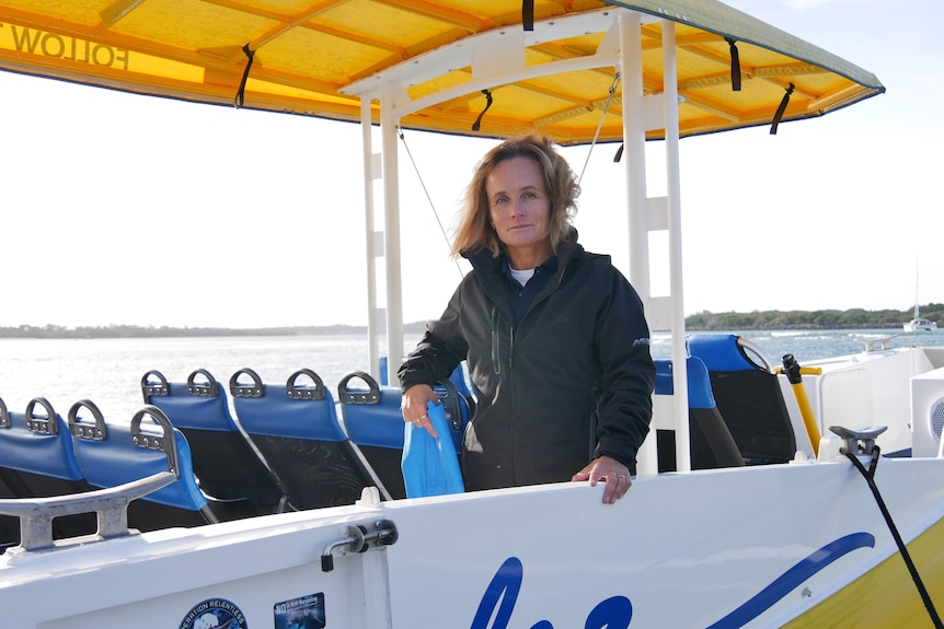A fair-haired, middle-aged woman wearing a dark, zip-up jacket is standing on a tourist boat 