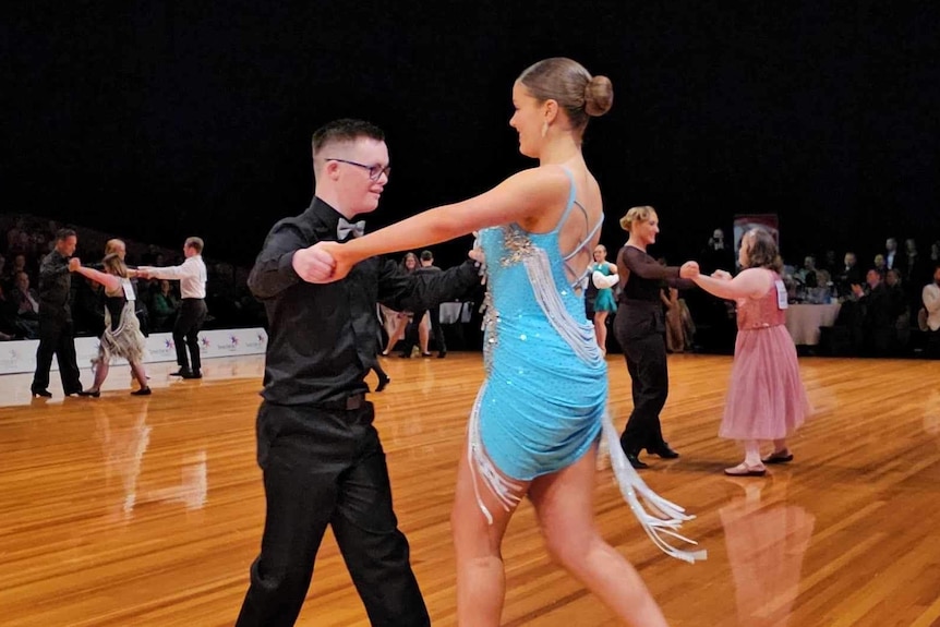 A young man and woman hold hands and dance on a hardwood dance floor surrounded by a crowd.