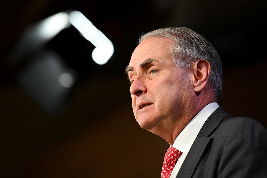 An older man in a suit and red, spotted tie, speaks into a microphone on a podium during an address.