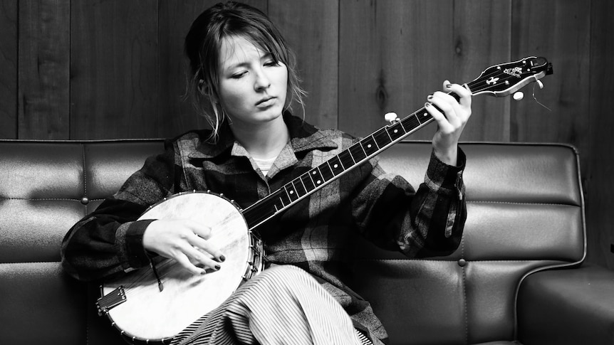 A black-and-white photo of Nora playing her banjo sitting down. She is wearing a checkered shirt and is concentrating.