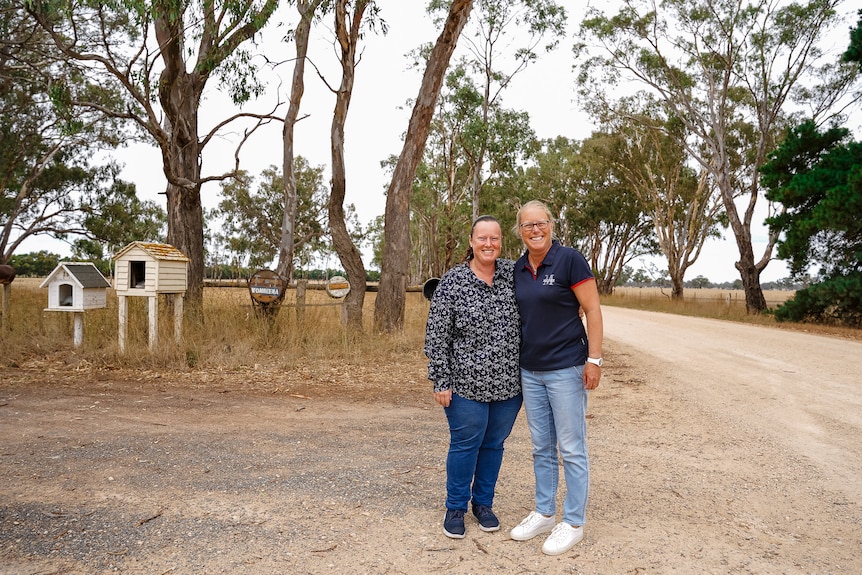 Two women in jeans and sneakers stand at a dirt crossroads smiling.