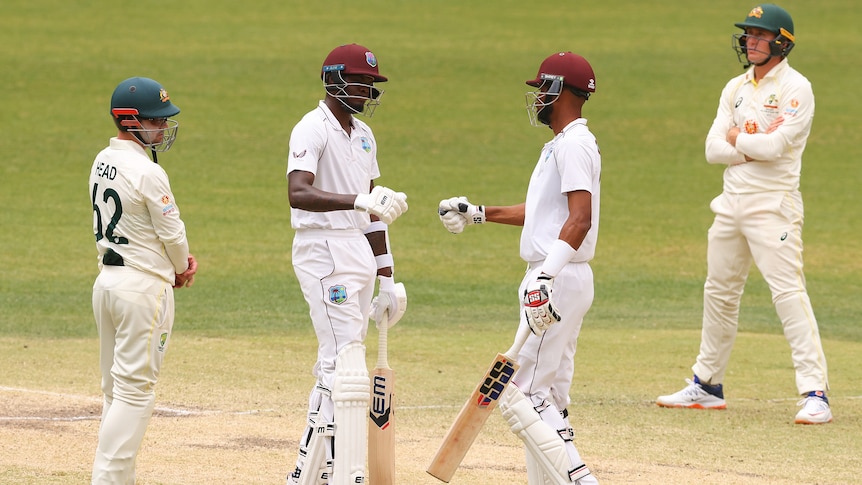 West Indies batters Alzarri Joseph and Roston Chase bump fists while Australian fielders look on during a Test match.