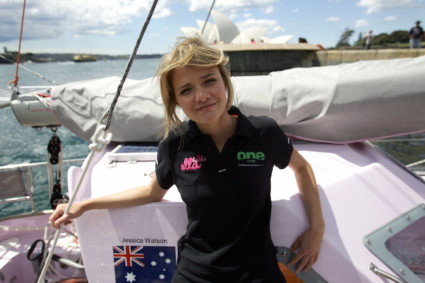 Jessica Watson stands smiling at the camera on her pink boat, leaning back, opera house and harbour in background.
