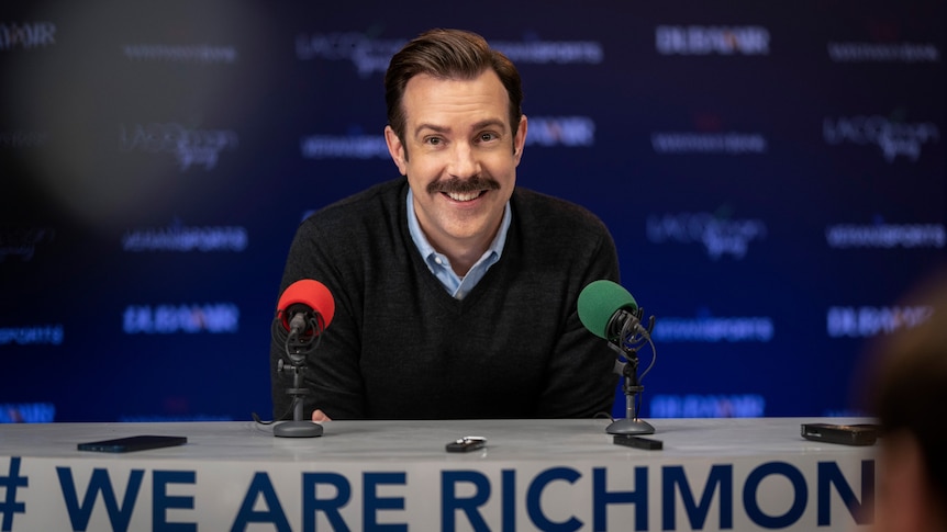 A 40-something man with a moustache smiles while seated at a table at a press conference. The tablecloth reads #WeAreRichmond.