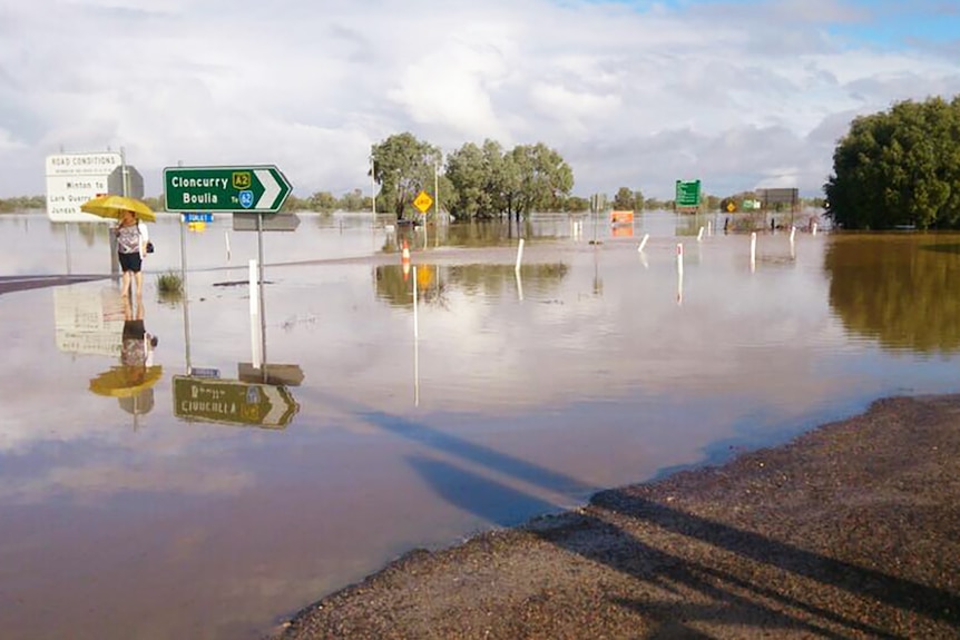 A road sign and a lady with an umbrella in a massive pool of water over the road