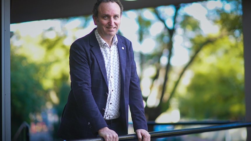 A man in a dark blazer and business shirt leaning with hands on railing