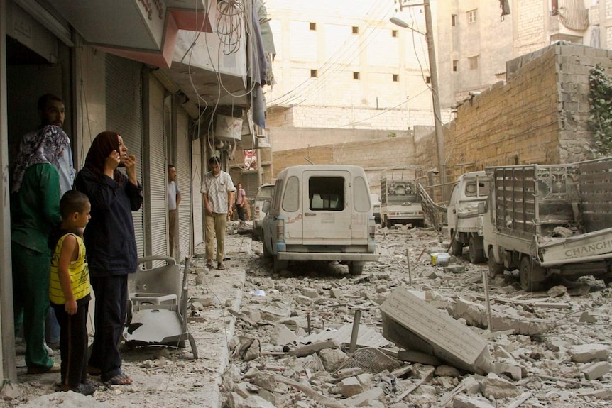 A street is strewn with bricks turned grey by dust as a crowd and woman adjusting her dark hijab look out onto the scene.