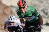 Caleb Ewan grimaces while cycling up a hill. He is at the front of a group of riders.