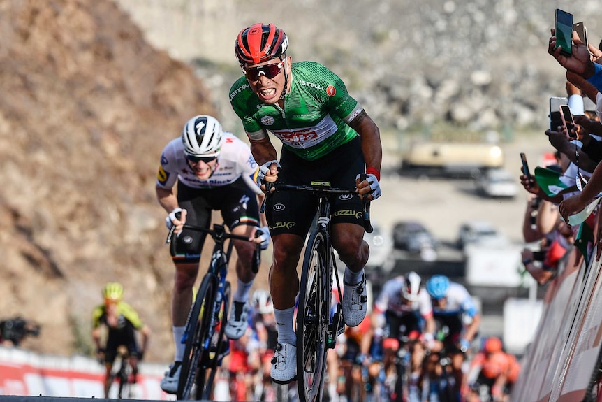 Caleb Ewan grimaces while cycling up a hill. He is at the front of a group of riders.