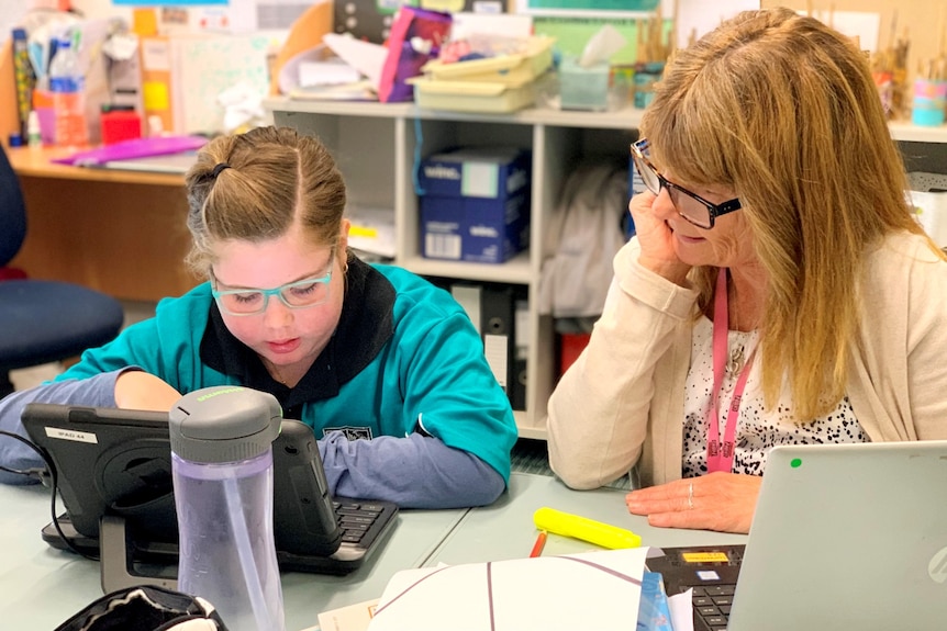 A young girl in a blue school shirt with glasses hovering over an ipad with a teacher looking over her shoulder while she works