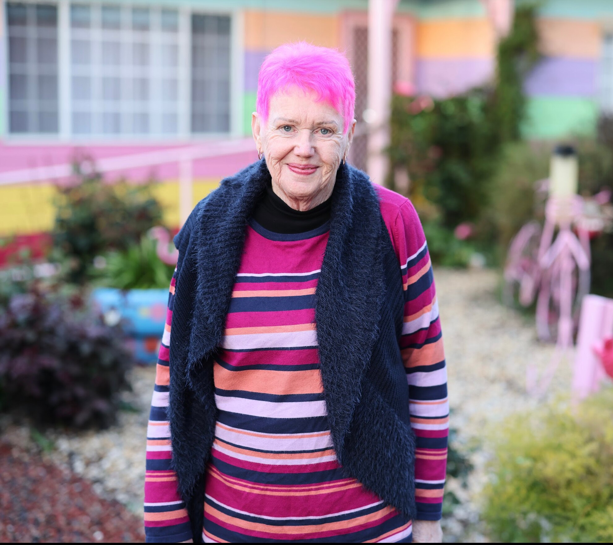 Gwenda Darling, a woman with pink hair, stands in a garden wearing a black scarf and striped jumper.