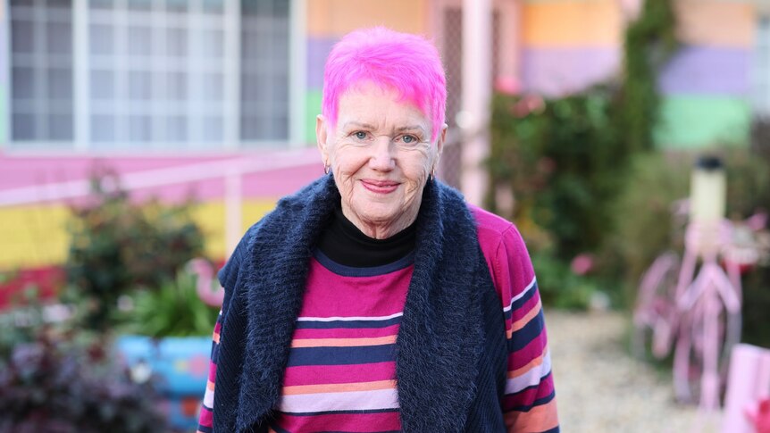 Gwenda Darling, a woman with pink hair, stands in a garden wearing a black scarf and striped jumper.