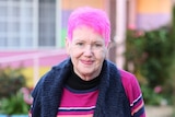 Gwenda Darling, a woman with pink hair, stands in a garden wearing a black scarf and striped jumper.