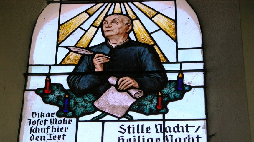 A stained glass image window featuring a saint-like man writing on a scroll with a feather.
