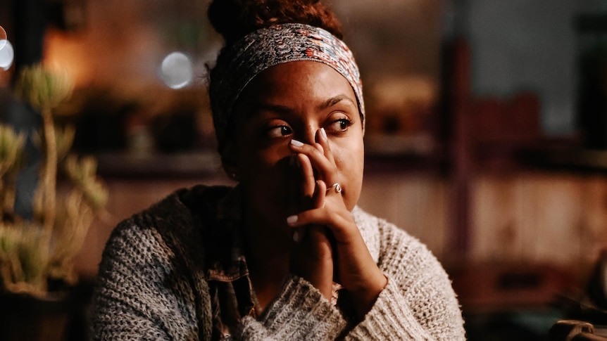 A woman on woolen jumper sits with elbows on a table with hands at mouth, staring off as if in thought. 