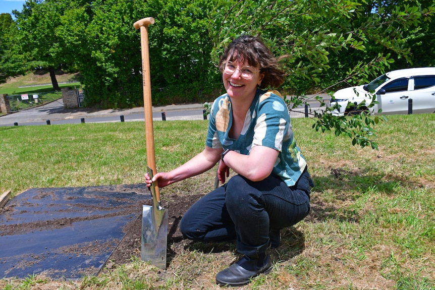 Woman with short hair and glasses crouches next to a shovel in a leafy park setting. 