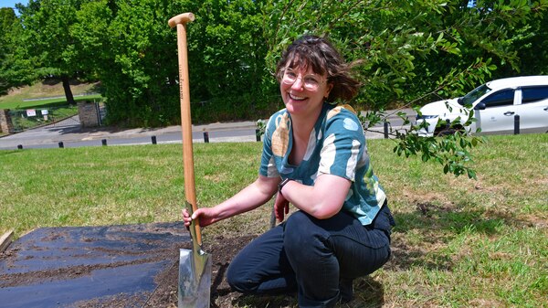 Woman with short hair and glasses crouches next to a shovel in a leafy park setting.