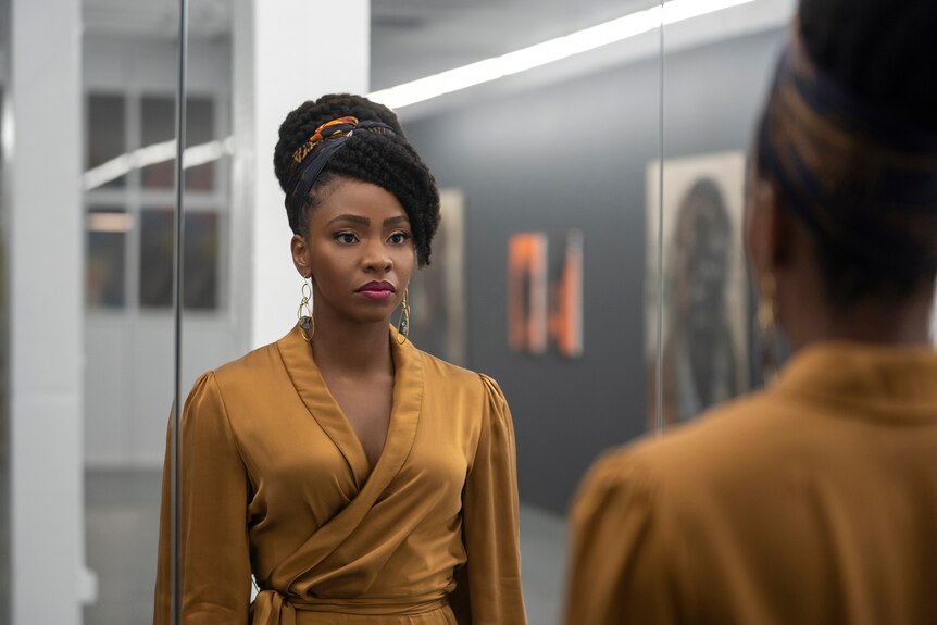 Teyonah Parris dressed in a tan silk wrap dress and dangling gold earrings stares somberly at her reflection in the mirror.