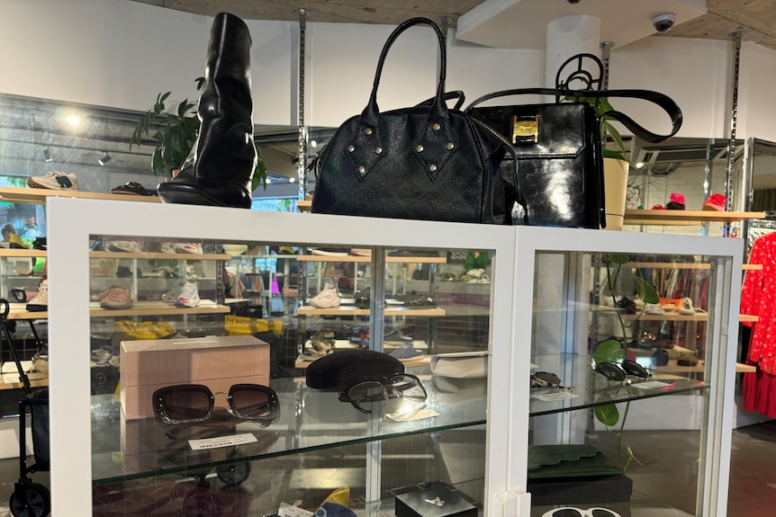 A glass cabinet inside a store with sunglasses for sale. Two black handbags and a pair of black boots are on top of the cabinet.