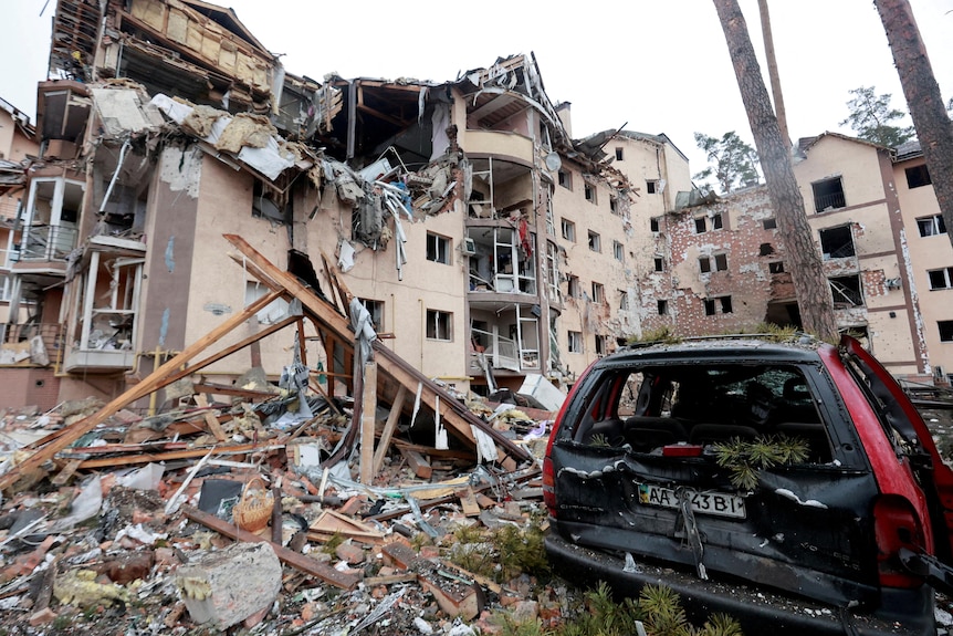 A building with wall gaping open, rubble on ground, destroyed car in foreground.