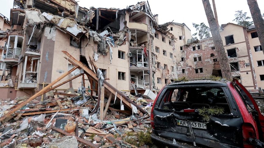 A building with wall gaping open, rubble on ground, destroyed car in foreground.