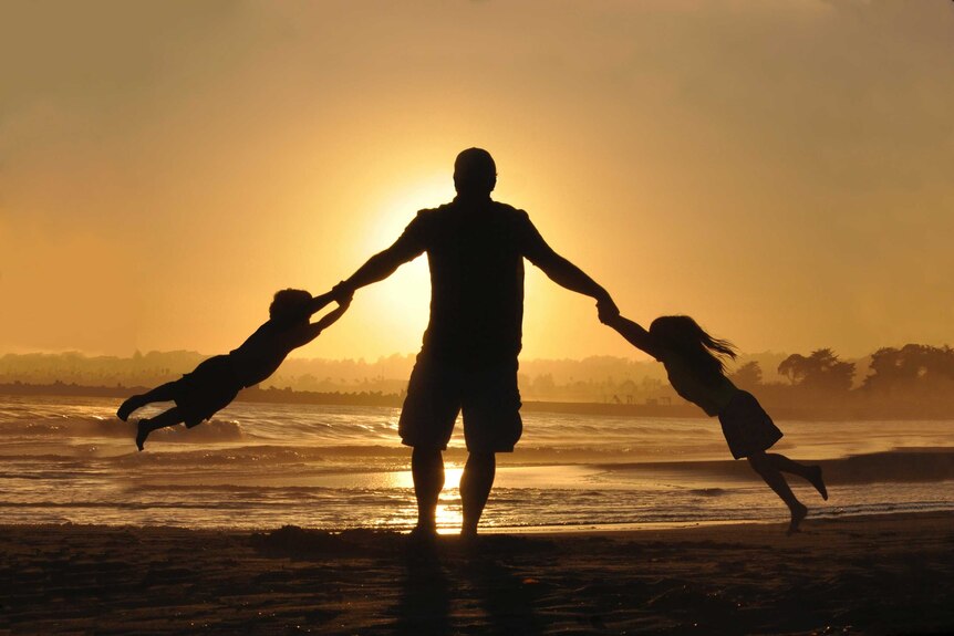 Silhouette of a man swinging two small children around on his arms on a beach at sunset.