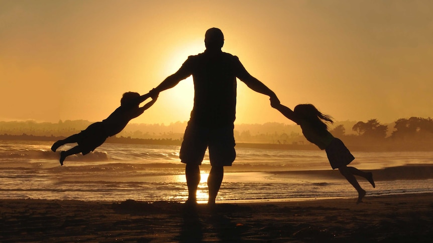 Silhouette of a man swinging two small children around on his arms on a beach at sunset.