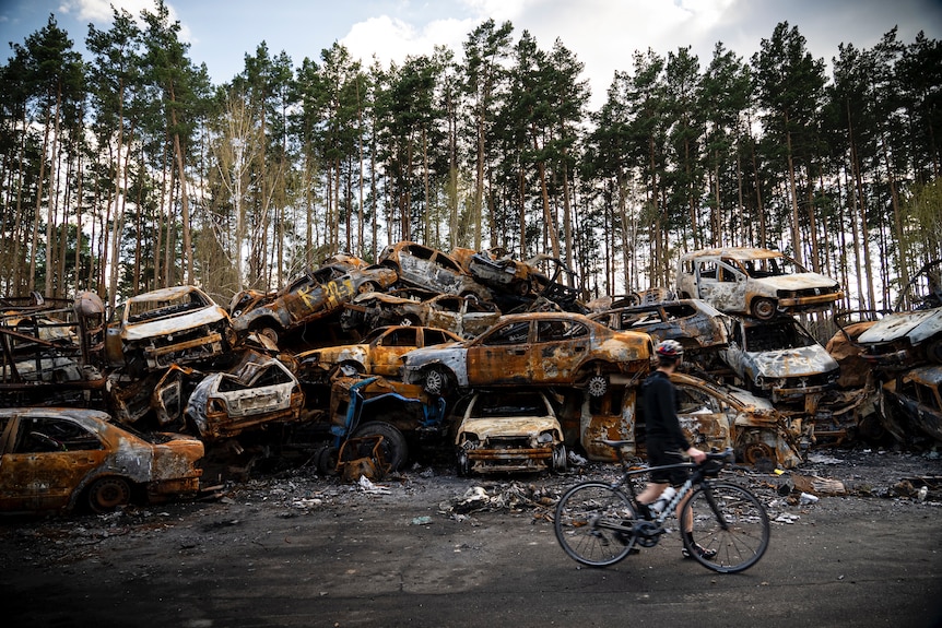 A man rides past a massive pile of burned out cars in a scene that looks like something out of Mad Max
