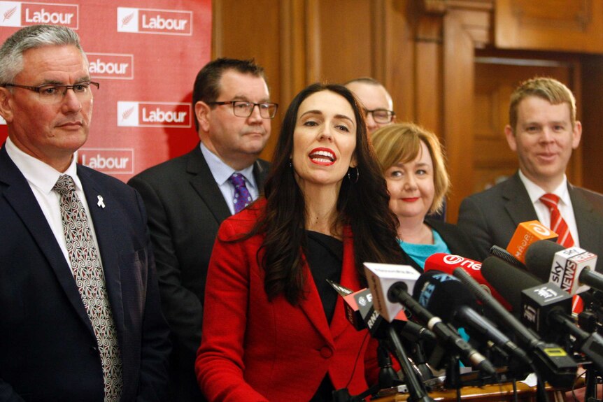 New Zealand Labour Party leader Jacinda Ardern speaks to the media, surrounded by several of her party colleagues.