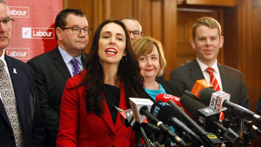 New Zealand Labour Party leader Jacinda Ardern speaks to the media, surrounded by several of her party colleagues.