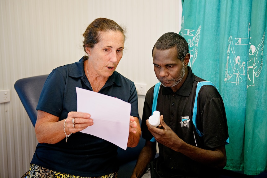 An Aboriginal man holds a small white device and looks at a piece of paper being shown to him by a woman. 