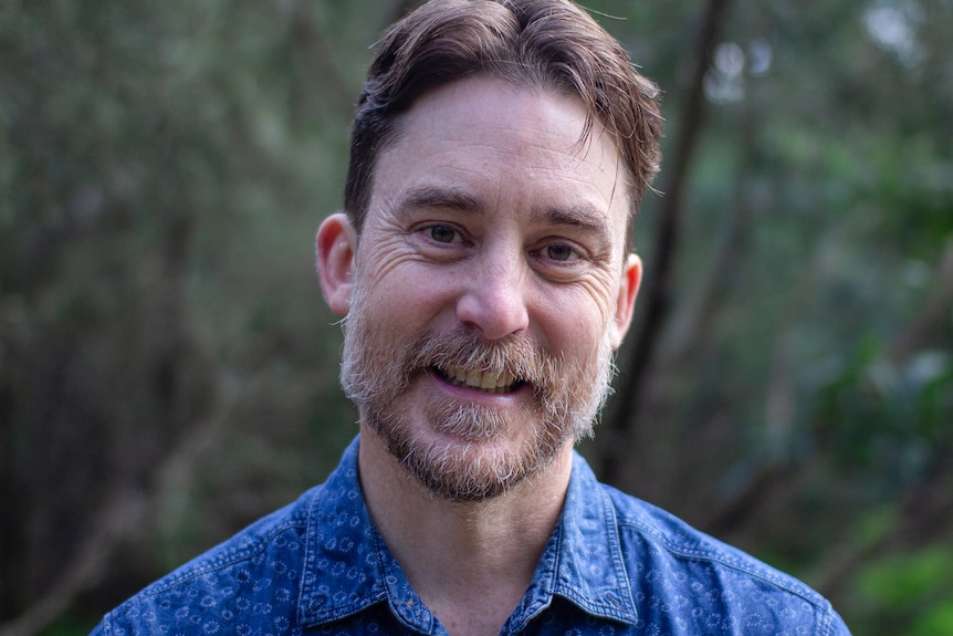 A close up of a man with brown hair and beard smiles at the camera, wearing a blue shirt. 