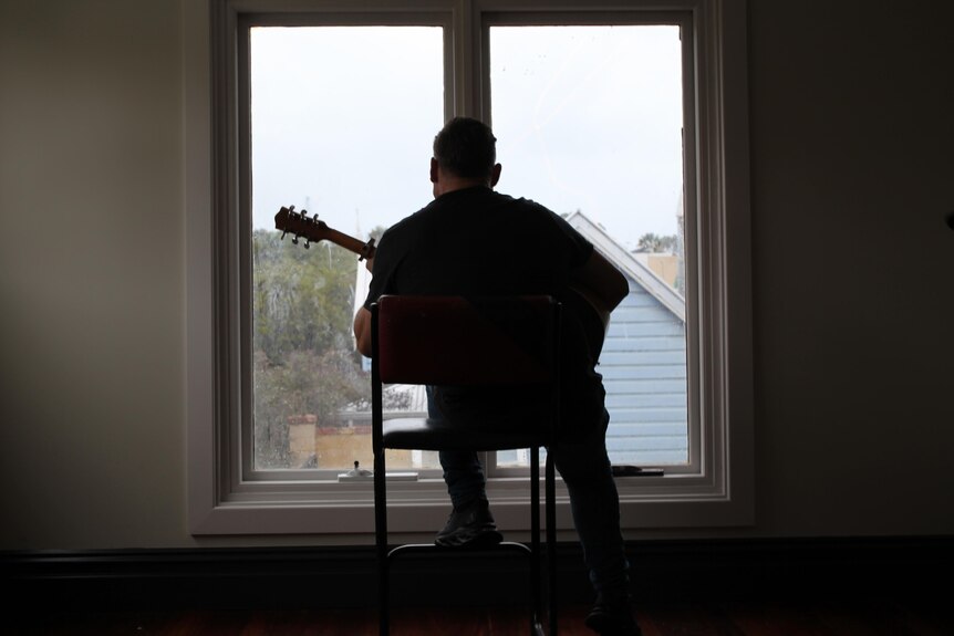 A man in a dark room plays a guitar in front of a window