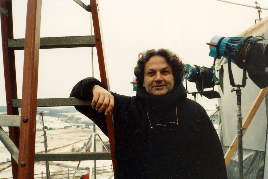 A man leans on a ladder, with film lights, and snow in the background 