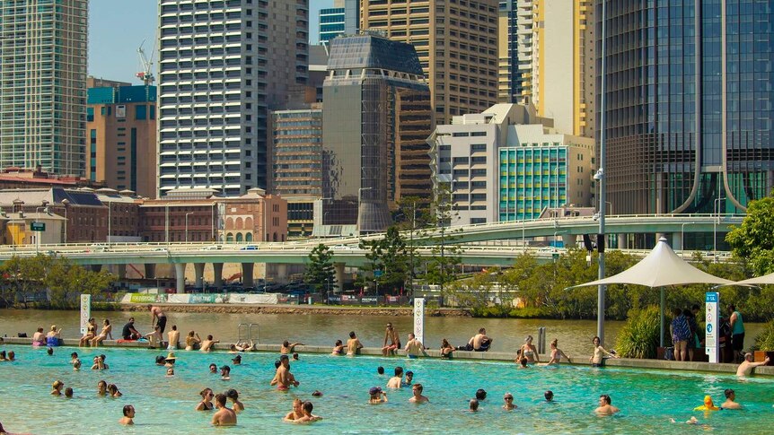 A crowd cools off in the South Bank lagoon, with Brisbane city skyscrapers in the background.