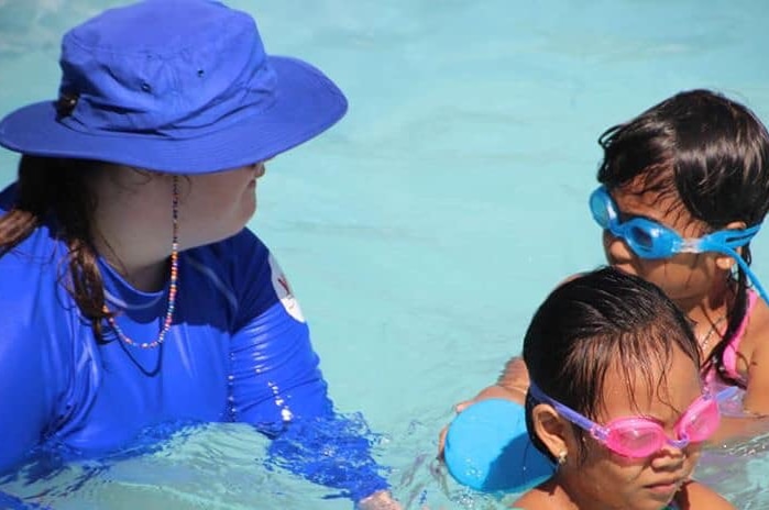 Woman in blue had on right of photo watches two young swimmers wearing goggles on left of photo.