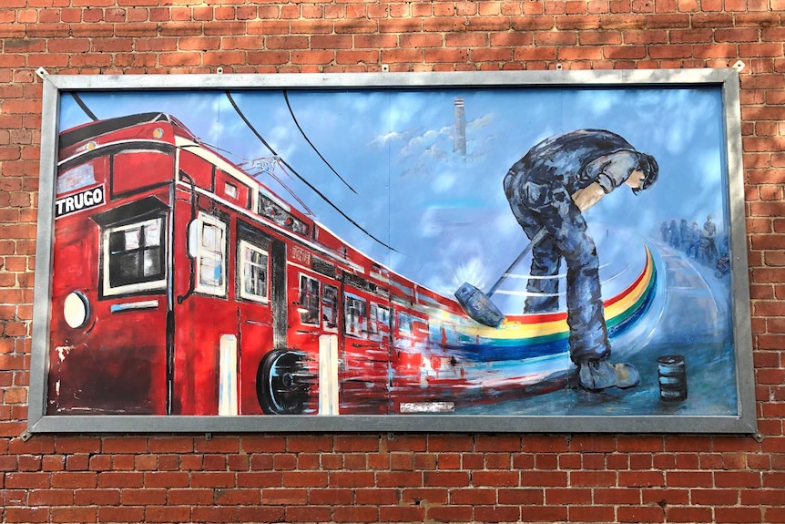 A mural showing a tram and a man playing trugo hangs on a red brick wall.