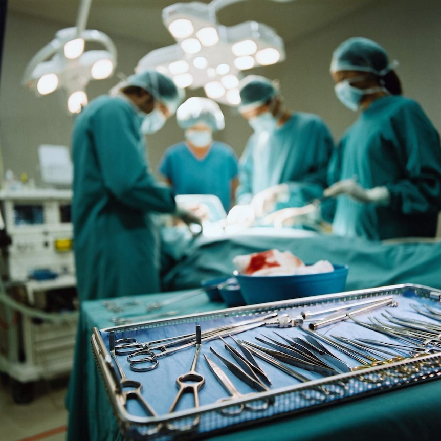 Four medical staff stand in the background, blurred, over an operating table. Medical instruments sits on a tray in foreground.