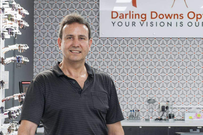 A man standing in front of a counter with a shelf of glasses to his left.