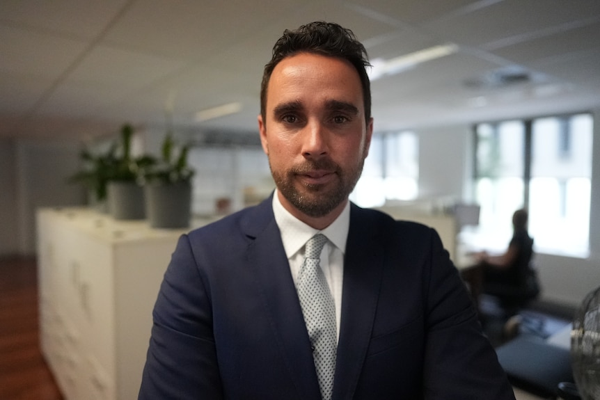 A young man with short hair in a suit poses for a photo in an office.