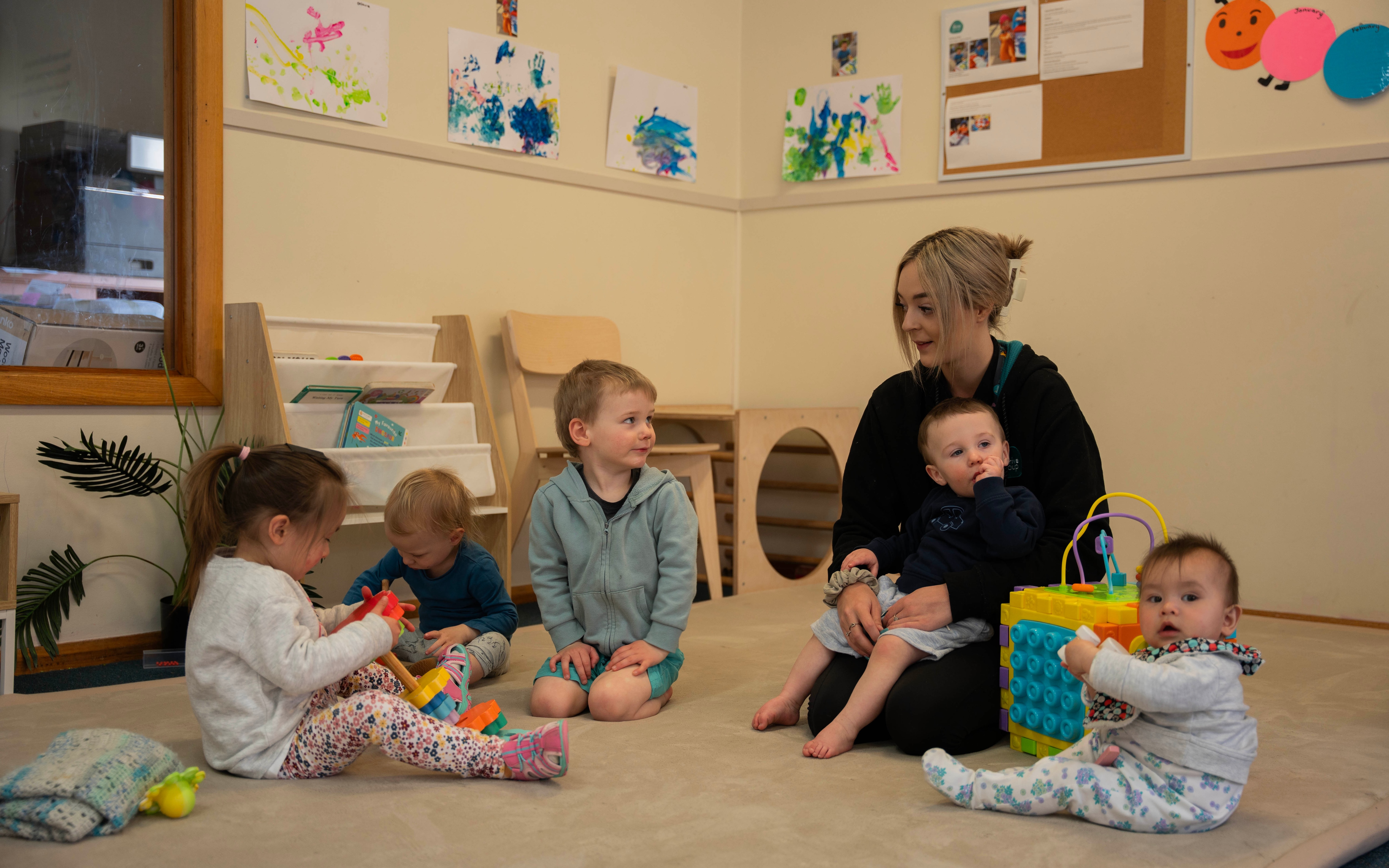 A woman sits on a mat with a babies and children playing nearby.