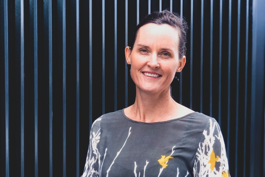 Smiling woman wears grey top with white and yellow print, dark hair tied back, stands in front of a blue paneled wall. 