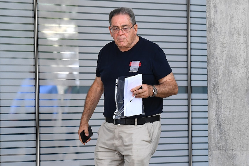 a man wearing a black t shirt and reading glasses walks out of a courthouse holding a clear folder with papers in it
