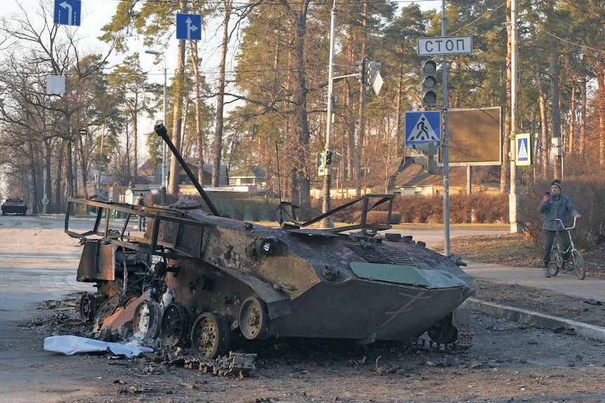 A Russian vehicle is charred and destroyed with twisted metal on the street, while a boy on a bike takes a photo
