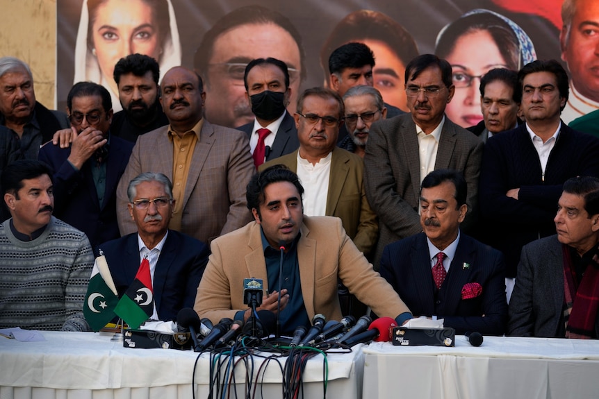 A young South Asian man in tan jacket speaks in front of a group at an indoor media conference