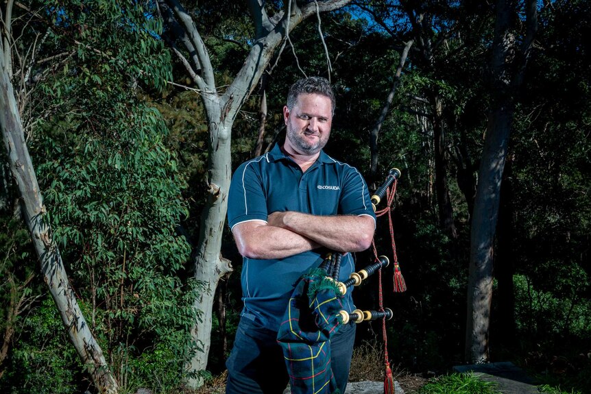 Jeremy holds his bagpipes while standing in front of gum trees.