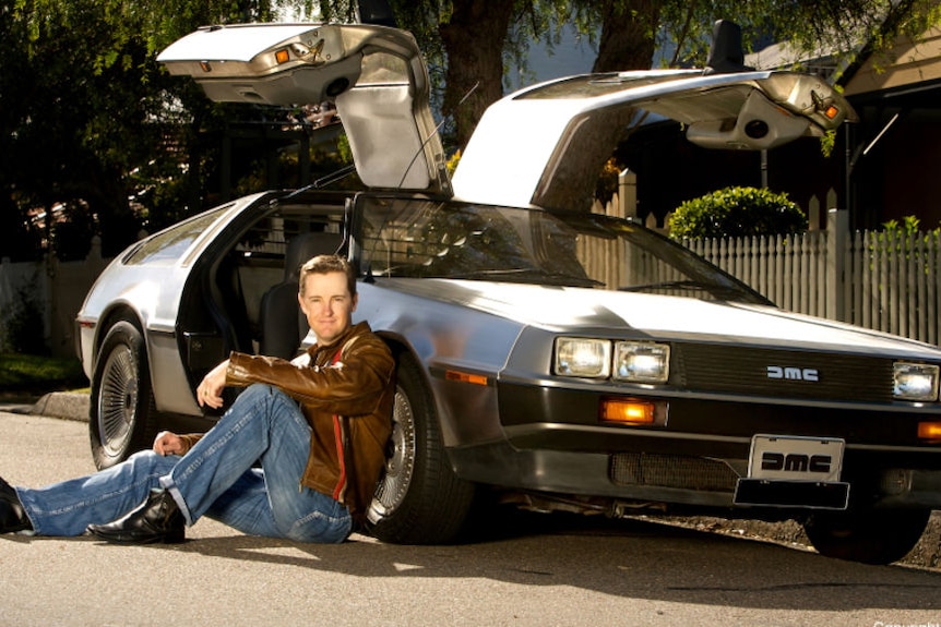 A man sits on the road leaning on his DeLorean car