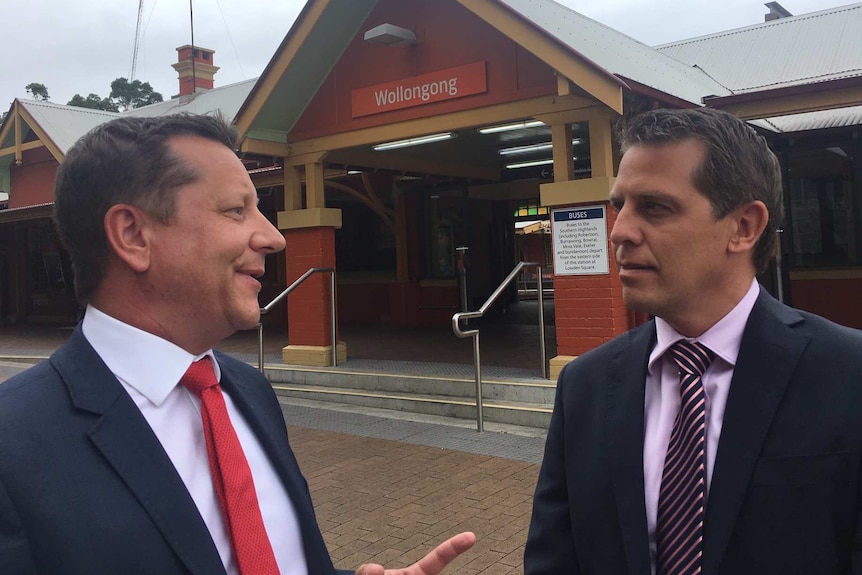 Two men in suits stand speaking outside a train station.